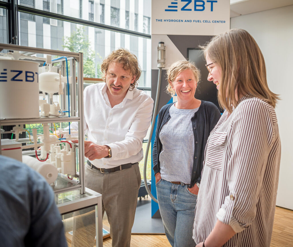 Wissenschaftler:innen besprechen sich neben dem Modell einer Cracker-Anlage zur Wasserstofferzeugung aus Ammoniak