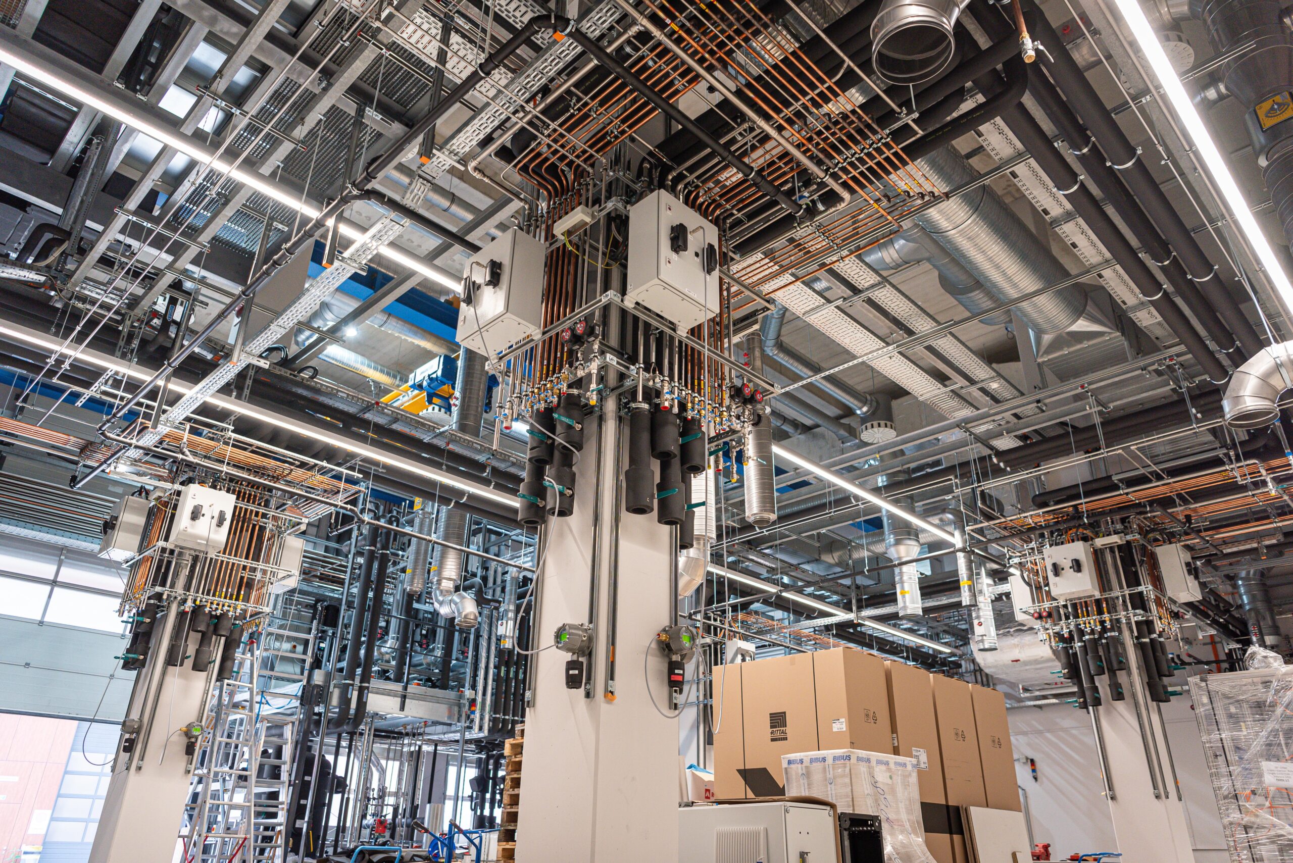 Das Hauptlabor hytechlab4nrw im ZBT. Zu sehen ist der Zustand des Labors im Aufbau. Das Bild zeigt vier tragende Säulen, dutzende Rohre und Leitungen, die Lüftungsanlage, mehrere Kartons und Maschinen im Aufbau.