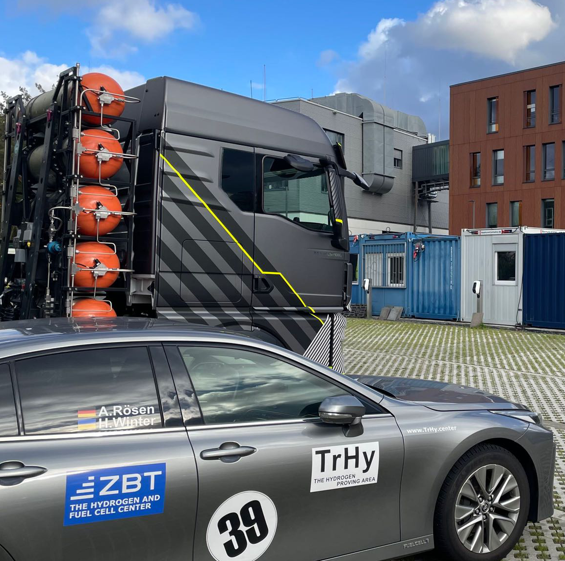 Eine Sattelzugmaschine mit Wasserstofftanks und ein brennstoffzellenbetriebener Pkw stehen auf dem Parkplatz vor den ZBT-Gebäuden.